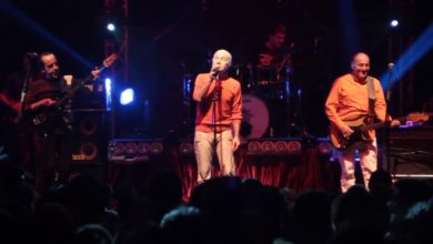 Three band members in orange tops perform on a lit stage with a drummer behind them, in front of a silhouetted crowd.
