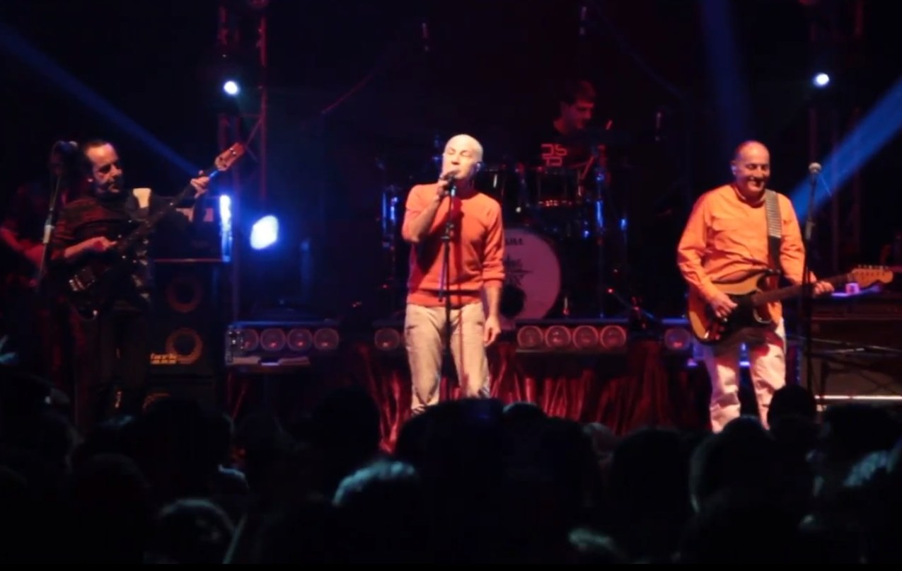Three band members in orange tops perform on a lit stage with a drummer behind them, in front of a silhouetted crowd.
