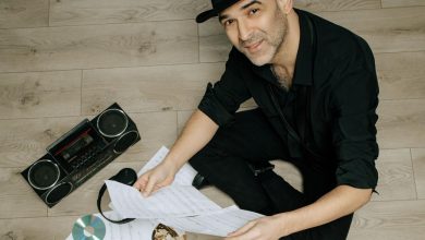 Man in a black hat and black clothes sits on a wooden floor holding sheet music, with a boombox, headphones and CDs beside him