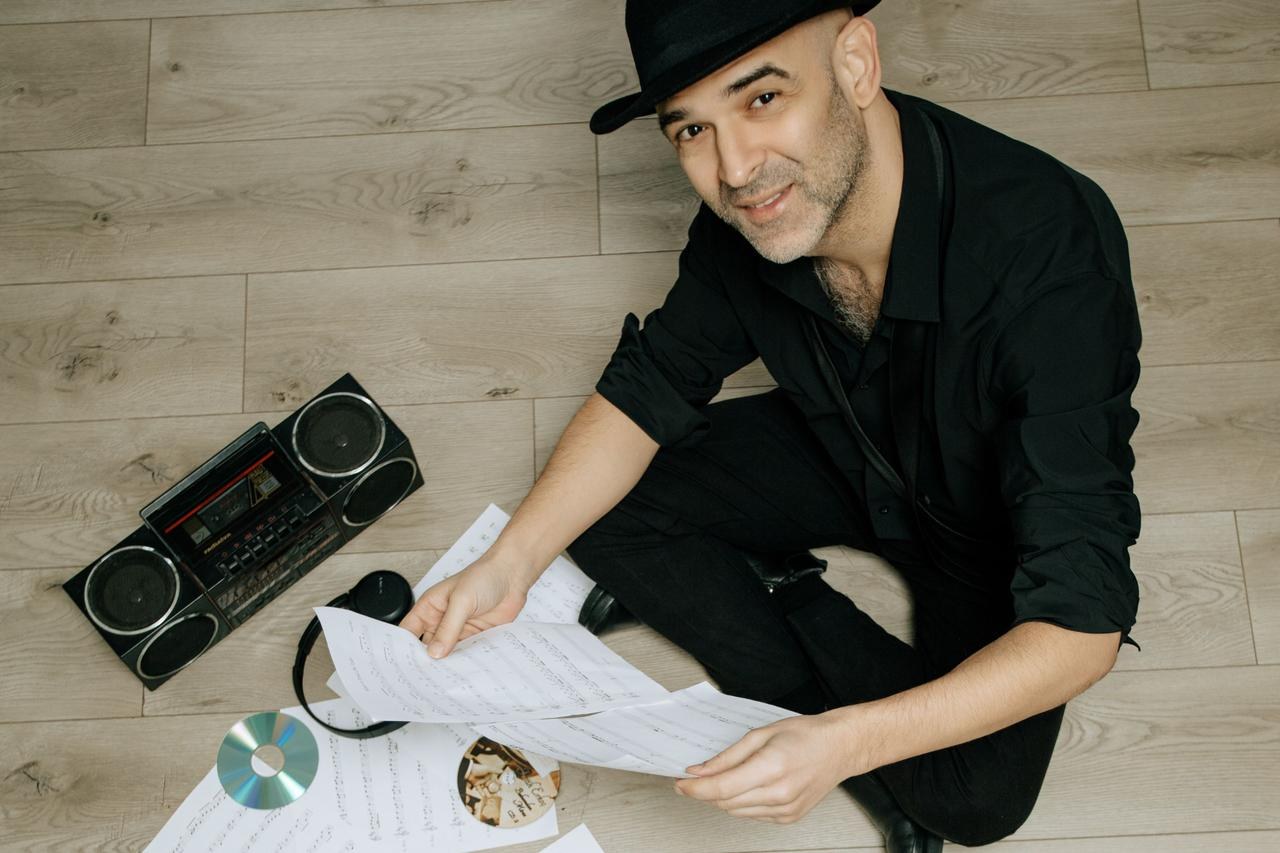 Man in a black hat and black clothes sits on a wooden floor holding sheet music, with a boombox, headphones and CDs beside him