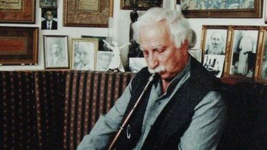 Elderly white-haired man with a mustache playing a reed flute (ney) on a sofa, in a room filled with framed portraits and calligraphy