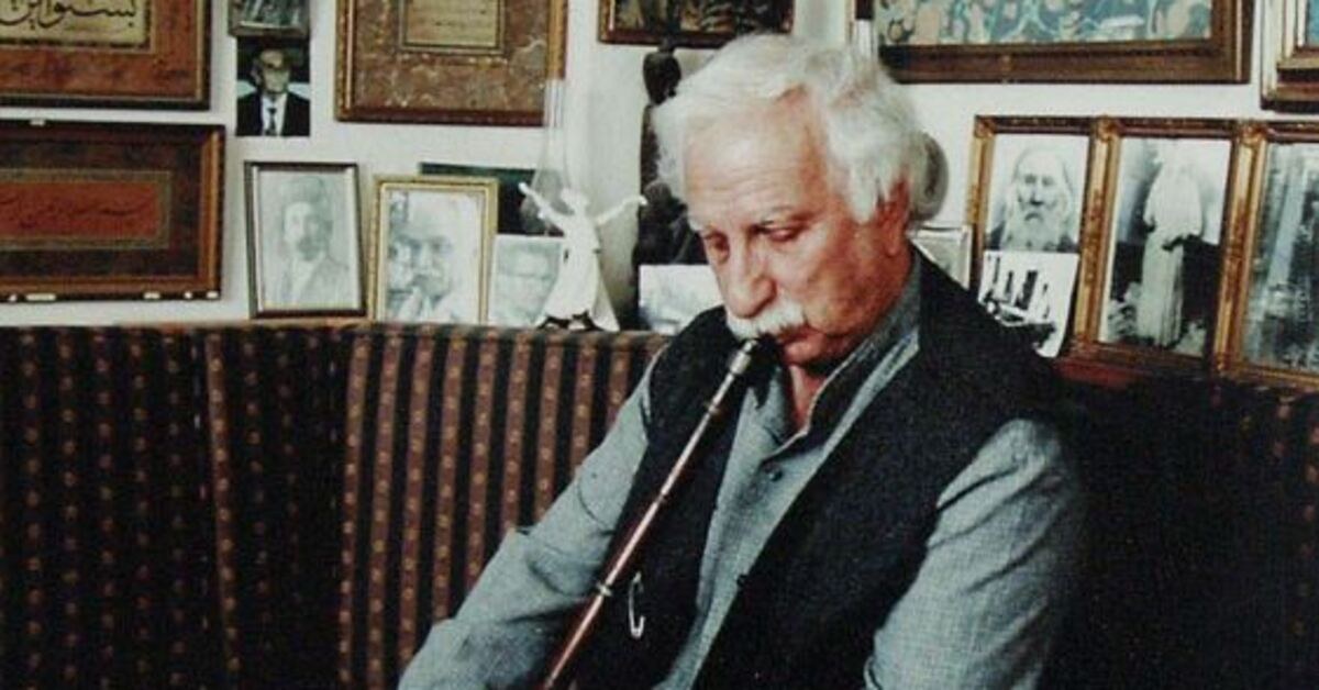 Elderly white-haired man with a mustache playing a reed flute (ney) on a sofa, in a room filled with framed portraits and calligraphy