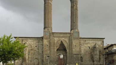 Twin-minaret stone madrasa with a tiled portal under a stormy sky, with people seated in the park in front, in Erzurum