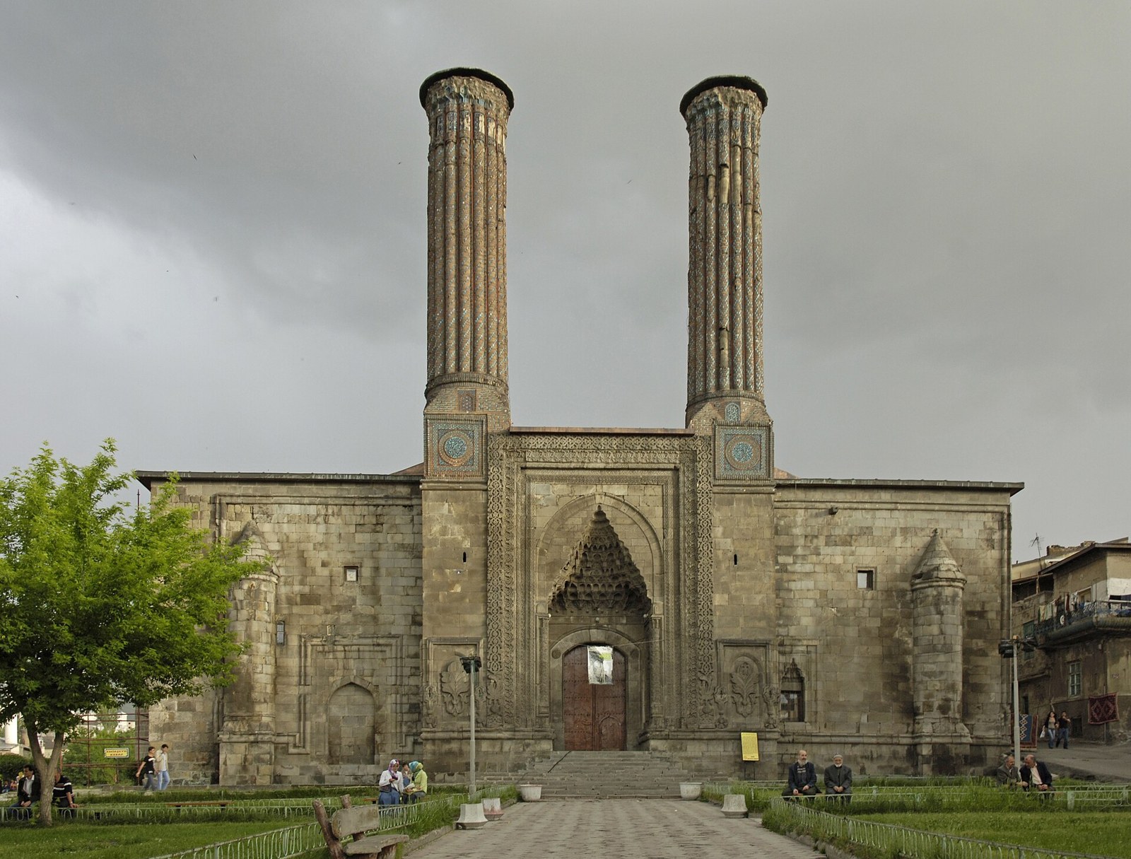Twin-minaret stone madrasa with a tiled portal under a stormy sky, with people seated in the park in front, in Erzurum