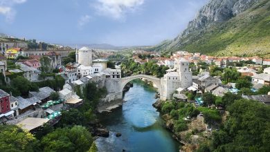 Ottoman stone bridge arching over a turquoise river in Mostar, with old stone houses, a minaret and mountains behind.
