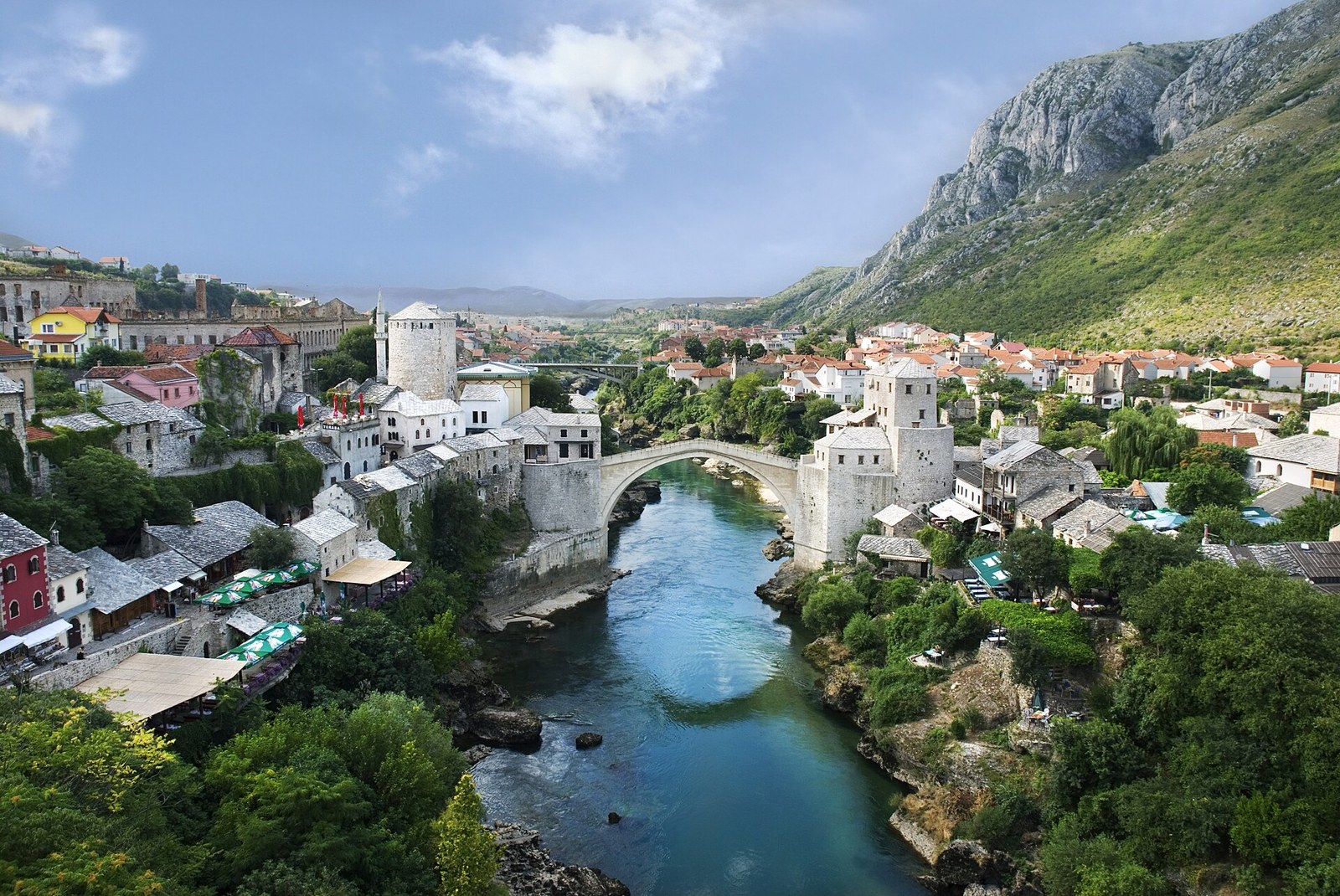 Ottoman stone bridge arching over a turquoise river in Mostar, with old stone houses, a minaret and mountains behind.