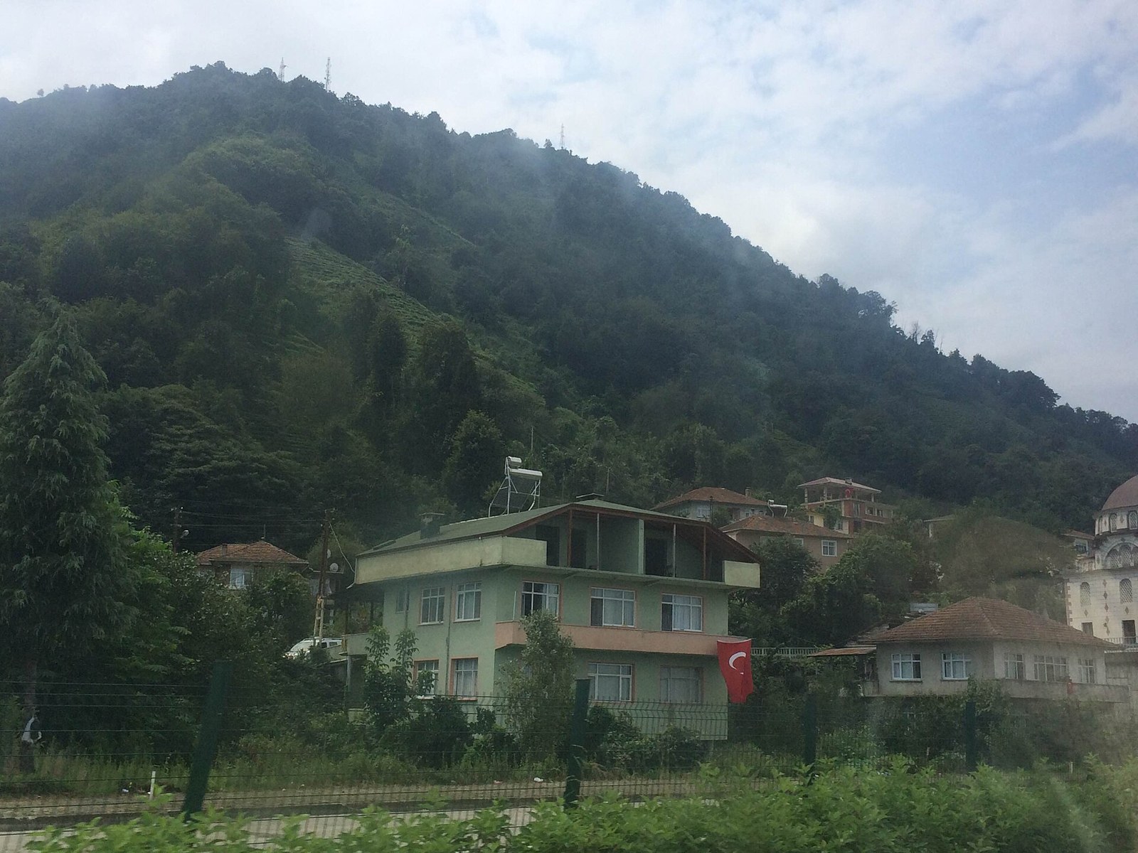 Forested Black Sea hillside with tea terraces above village houses and a Turkish flag hanging from a green building
