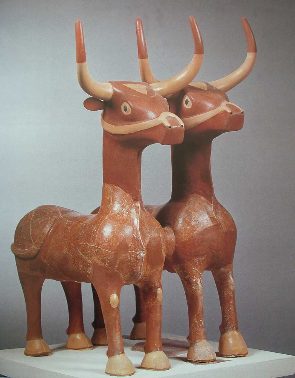 Pair of ancient Hittite-style terracotta bull statues with long curved horns on a white display plinth