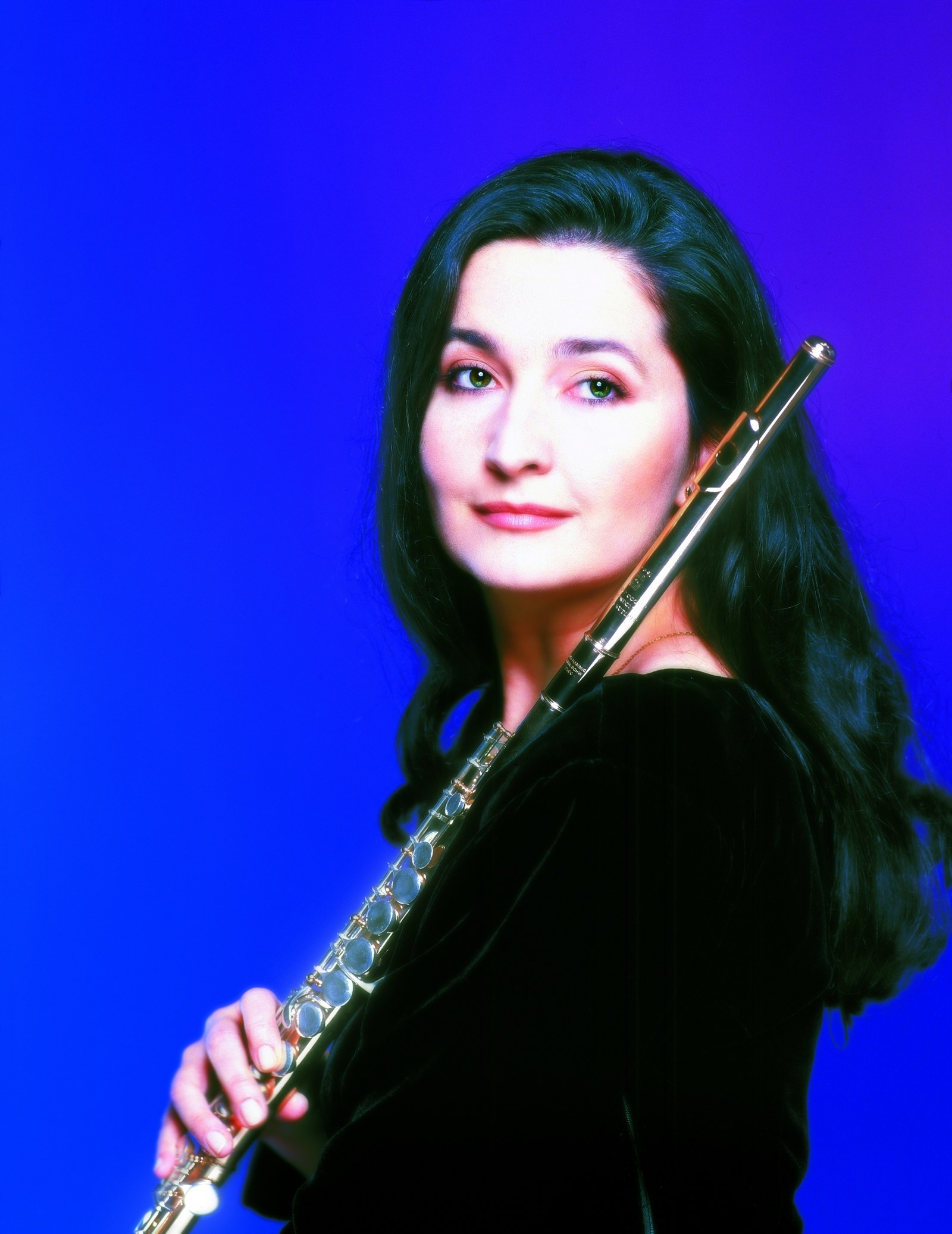 Studio portrait of a dark-haired woman in a black velvet top holding a flute against a deep blue background