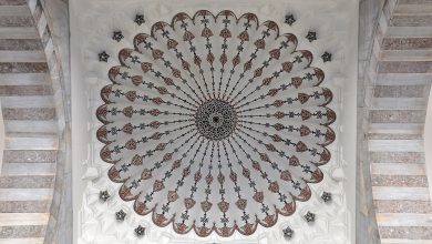 Looking up at an ornate white dome with red and black radial patterns, a lattice medallion at its center and striped marble arches.