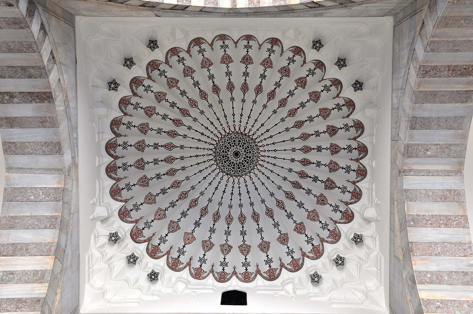 Looking up at an ornate white dome with red and black radial patterns, a lattice medallion at its center and striped marble arches.