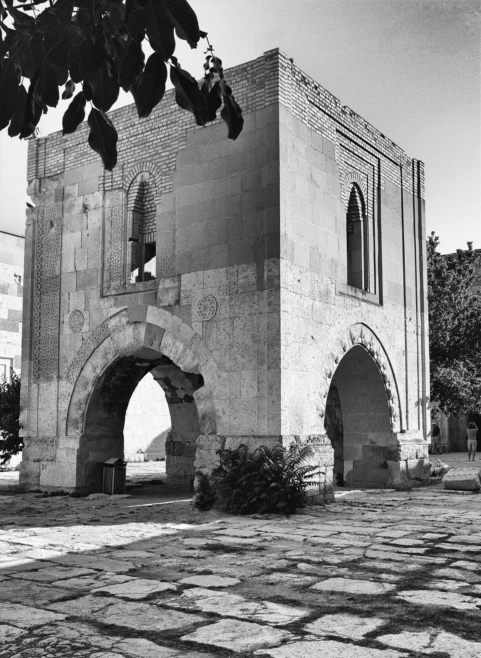 Black-and-white view of the stone mosque pavilion on arches in the courtyard of Sultanhanı Caravanserai, with paving and tree leaves.