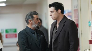 Two men in dark suits talk in a hospital corridor, a gray-haired bearded older man facing a younger dark-haired man.