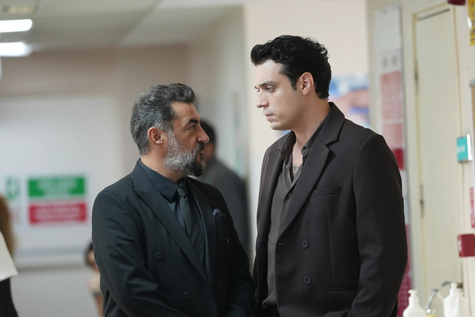 Two men in dark suits talk in a hospital corridor, a gray-haired bearded older man facing a younger dark-haired man.