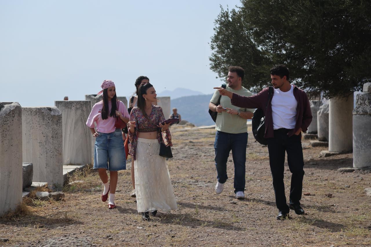 Four young people walk and talk among ancient stone columns at a hilltop ruin site under a hazy sky.