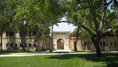 Stone gateway and striped masonry wall of a historic building behind shady plane trees and a lawn in a palace garden