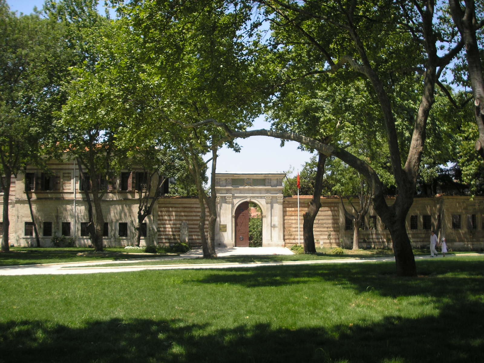 Stone gateway and striped masonry wall of a historic building behind shady plane trees and a lawn in a palace garden