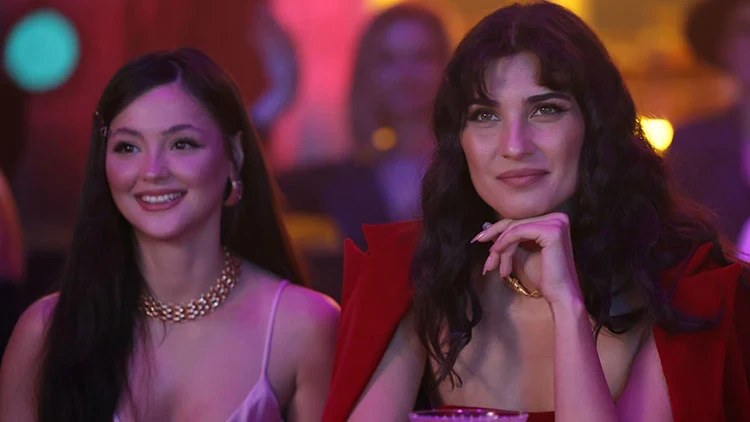 Tuba Büyüküstün and another woman smiling in a dimly lit setting, possibly a scene from the film 'Sultana'.