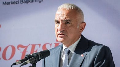 A gray-haired man in a dark suit and tie speaks at microphones in front of a banner with red lettering.