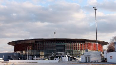 Exterior of a large circular indoor arena with a red-and-glass facade and wide roof, seen from an empty parking lot under cloudy skies