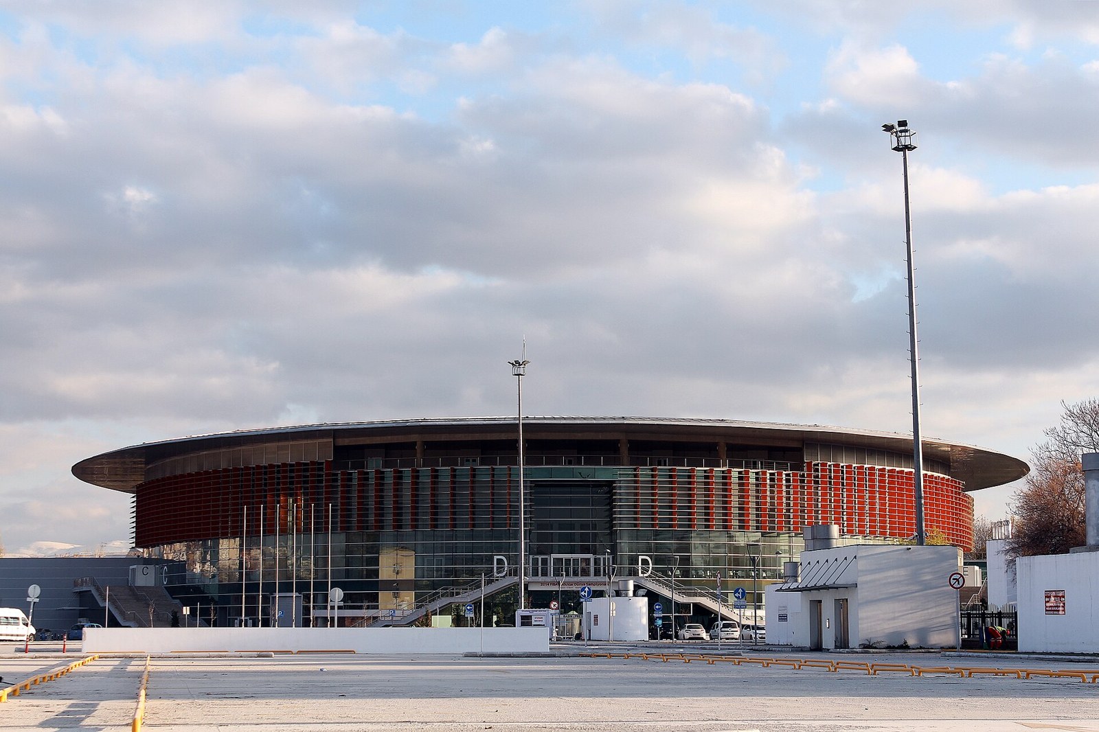 Exterior of a large circular indoor arena with a red-and-glass facade and wide roof, seen from an empty parking lot under cloudy skies