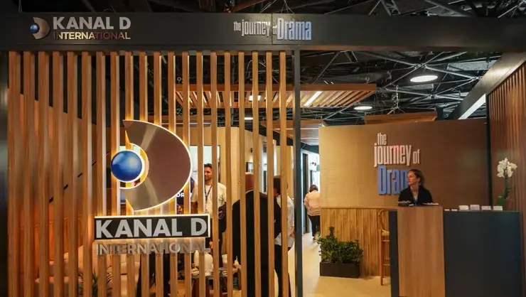 Kanal D International stand with wooden slats and 'The Journey of Drama' signs, with visitors and a staff member at a desk