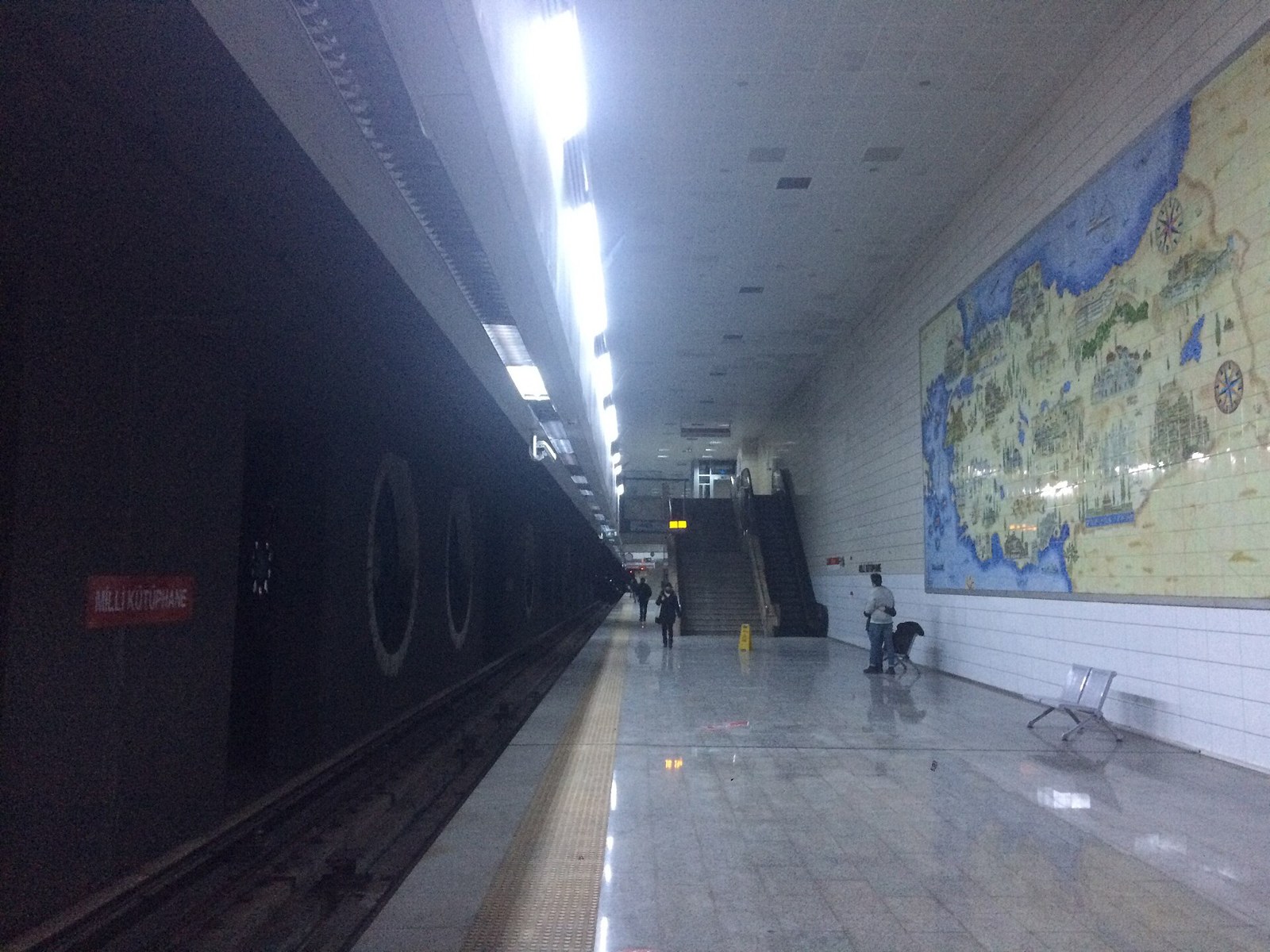Metro platform at Milli Kütüphane station with a tiled map mural of Türkiye on the wall and a few passengers walking.