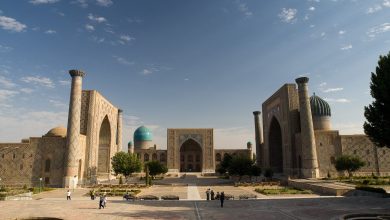 Wide view of a historic Central Asian square with three tiled madrasas, minarets and turquoise domes under a blue sky