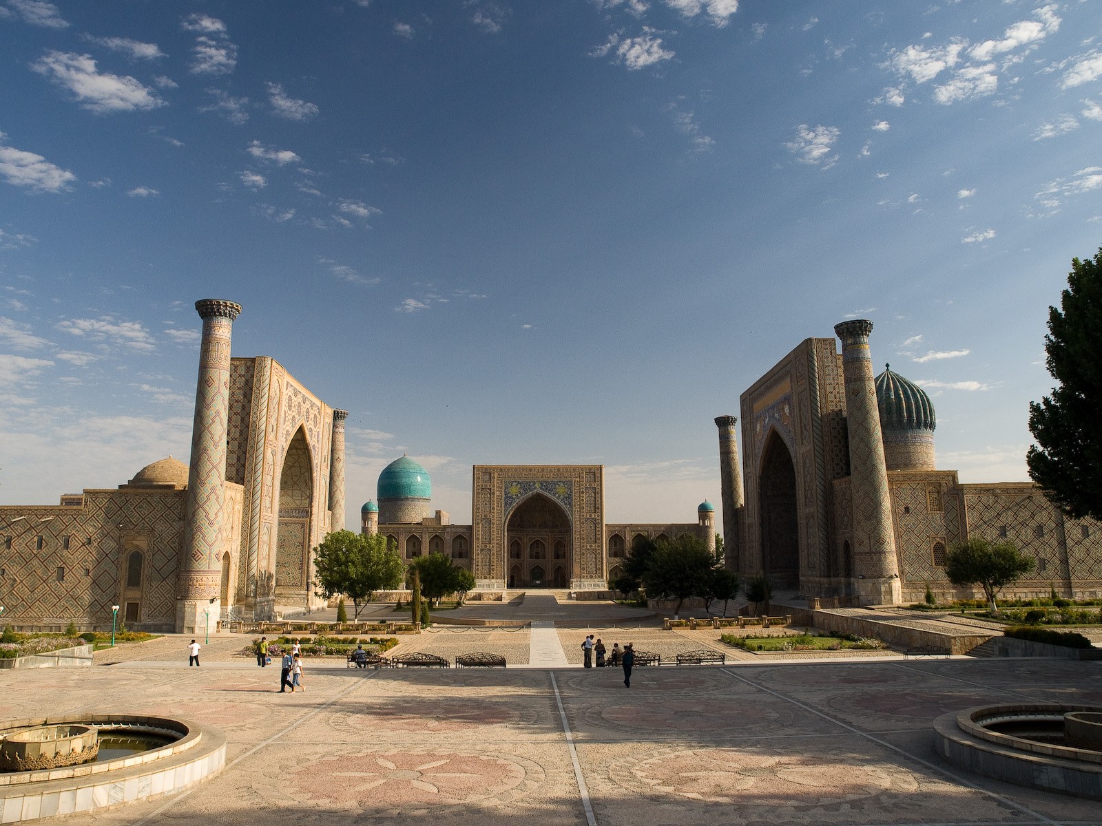 Wide view of a historic Central Asian square with three tiled madrasas, minarets and turquoise domes under a blue sky