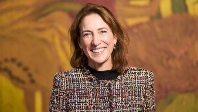 Smiling woman in a colorful tweed jacket stands in front of a large yellow and orange painting.