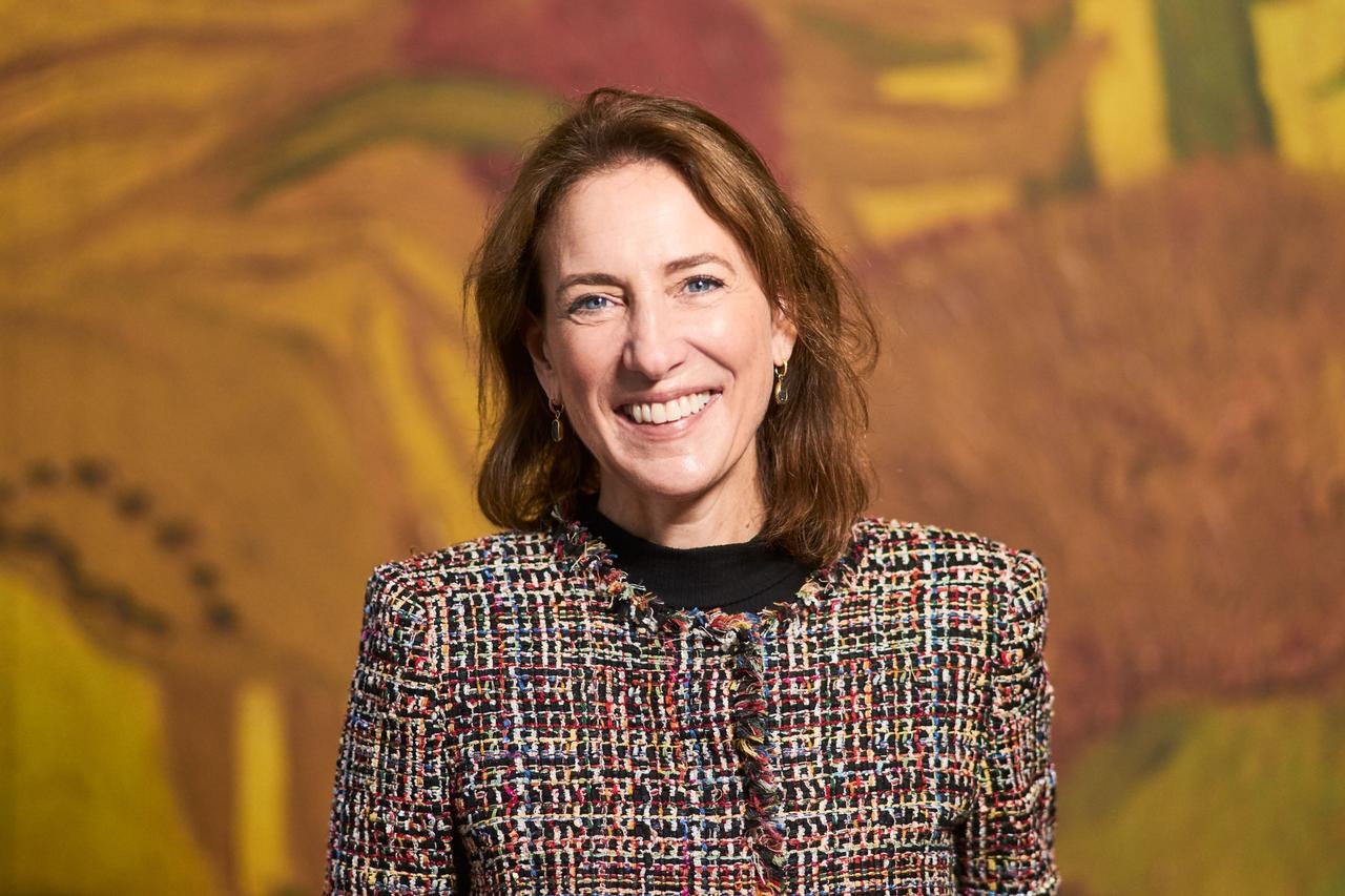 Smiling woman in a colorful tweed jacket stands in front of a large yellow and orange painting.